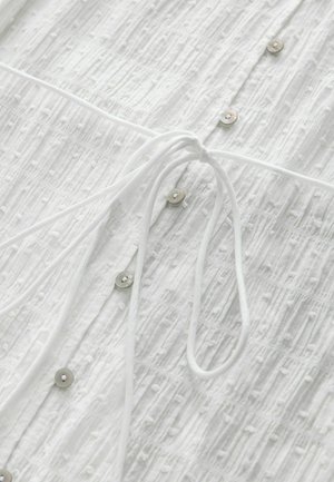 Weiße Bluse mit strukturiertem Stoff und erhabenen Mustern, silbernen Knöpfen und einer gebundenen Kordel am Ausschnitt.