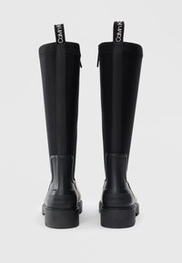 Bottes noires en caoutchouc imperméables, arrivant au genou, avec une base en caoutchouc lisse, une tige texturée et des accents de logo sur les languettes de traction. Fini lisse, semelle robuste.