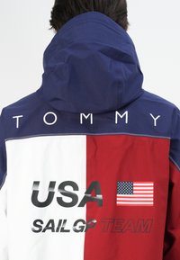 Person iført en blå, hvid og rød jakke med "TOMMY USA SAIL GP TEAM" og et USA-flag på ryggen.
