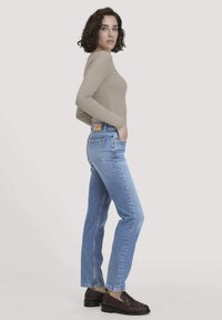 Nainen, jolla on kihara tumma hius, pukeutuneena vaaleanbeigeen pitkähihaisiin paitoihin, vaaleansinisiin suoriin farkkuihin ja tummanruskeisiin loafereihin, seisoo kyljellään.