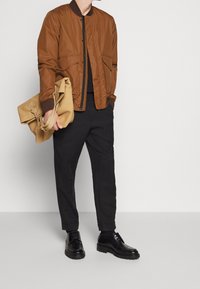 Brun bomberjacka med ribbad krage, svarta byxor och svarta skor. Håller en beige, strukturerad axelväska. Enkelt och modernt design.