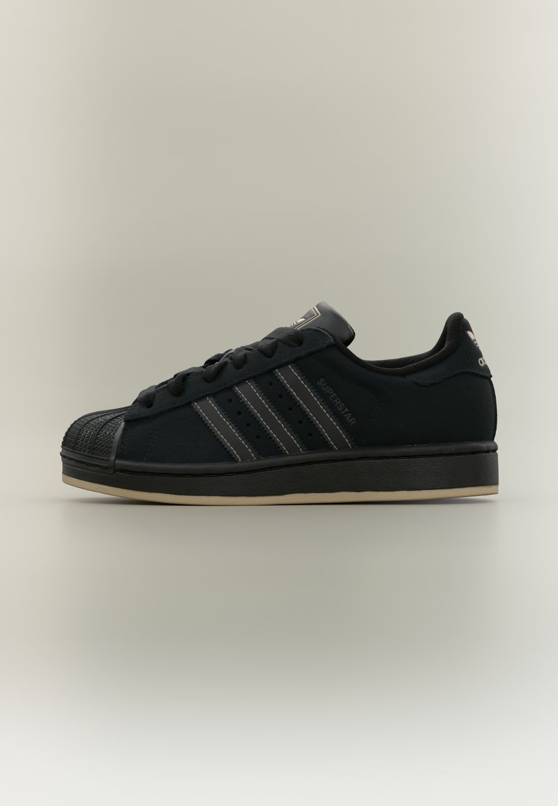 Μαύρο αθλητικό παπούτσι Adidas Superstar με υφή στο κεφαλόδεσμο, τρεις χαρακτηριστικές λωρίδες στο πλάι και μπεζ σόλα σε ουδέτερο φόντο.