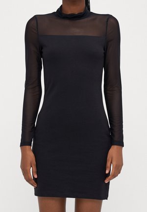 Woman wearing a black long-sleeve dress with sheer mesh upper chest and sleeves, standing against a plain white background.