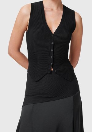 Femme portant un gilet noir sans manches côtelé boutonné sur un haut assorti côtelé et une jupe noire avec un ourlet asymétrique.