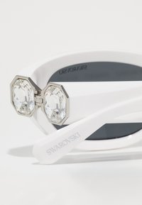 Vita solglasögon med en slank design, som har oktagonala kristalldetaljer och en glänsande yta. Graverad "SWAROVSKI" på skalmen.