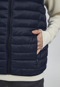 Mână în buzunarul lateral al unei veste matlasate bleu închis purtată peste o cămașă crem cu mânecă lungă și pantaloni închiși la culoare.