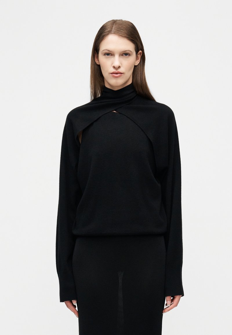 Γυναίκα με ίσια καστανά μαλλιά φορ άει μαύρο μακρυμάνικο τοπ με μοναδικό άνοιγμα και ψηλό λαιμόκοψιμο σε απλό λευκό φόντο.