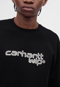 Gros plan d'une personne portant un sweat-shirt noir avec un texte brodé blanc "carhartt wip" sur la poitrine.