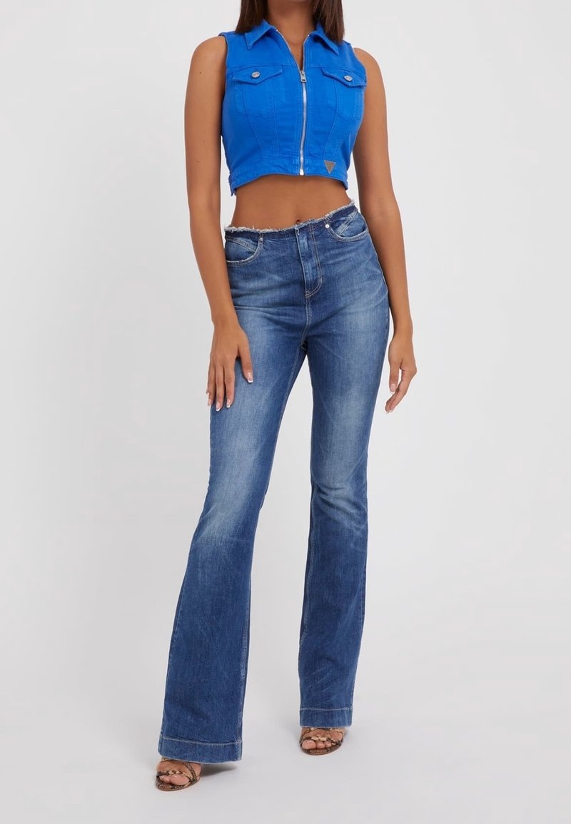 Femme portant un top court sans manches zippé bleu vif et un jean évasé bleu taille haute avec des sandales à talons, debout sur fond blanc.