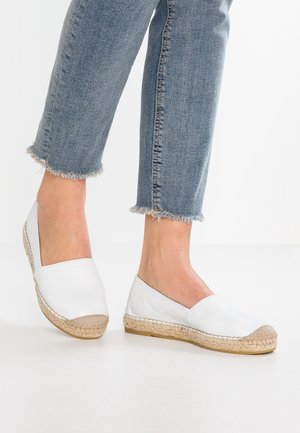 Person iført lyseblå jeans med flossede kanter og hvide espadrille-flade sko med vævede jutesåler på en ensfarvet hvid baggrund.