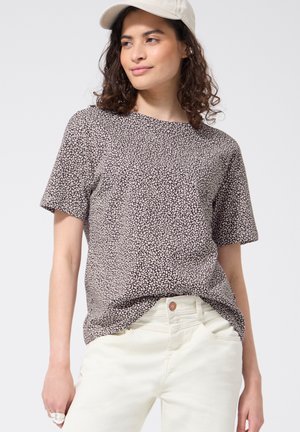 Femme portant une casquette beige, un haut à manches courtes imprimé léopard et un pantalon taille haute blanc cassé, debout devant un fond uni.