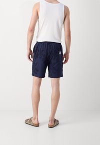 Uomo in canotta bianca senza maniche, pantaloni corti blu navy a motivo e sandali beige, in piedi di spalle su uno sfondo bianco uniforme.