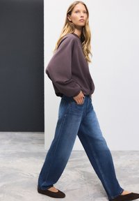 Oversized lilla sweatshirt, blå vidbenede jeans og brune flade sko. Glat stof med blød tekstur og afslappet pasform, vist mod en minimalistisk baggrund.