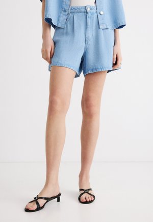 Woman wearing light blue denim shorts, matching denim jacket, and black low-heeled sandals standing on a white floor.