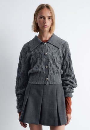 Gray knitted cardigan with a cable pattern, large collar, and decorative buttons; paired with a pleated gray skirt.
