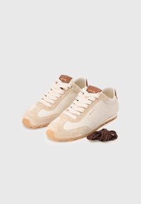 Adidași bej deschis, fabricați din suede și piele, cu detalii perforate. Prezintă șireturi crem și un accent pe toc de culoare maro. Șireturile maro sunt incluse.