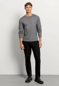 Grauer Pullover aus weichem Material, mit Rundhalsausschnitt und gerippten Bündchen, kombiniert mit schwarzen Hosen und schwarzen Schuhen.