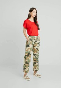 Woman with long dark hair wears red short-sleeve shirt, camo-pattern wide-leg pants, and beige sneakers, standing with hands in pockets.