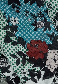 Tissu à motifs floraux avec des fleurs rouges, blanches et noires sur un fond géométrique bleu et vert. Matériau léger et texturé.