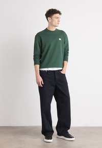 Grønn crewneck genser med en liten logo, parret med mørkeblå løse jeans og svarte joggesko med hvite såler.