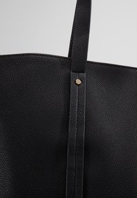 Sac en cuir noir avec une finition texturée, anses plates et un accent doré sur la couture latérale. Design minimaliste, sans fermeture visible.