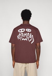 Uomo che indossa una maglietta marrone con occhi astratti bianchi e la grafica "KARMA 89" sul retro, pantaloni beige, in piedi contro uno sfondo semplice.