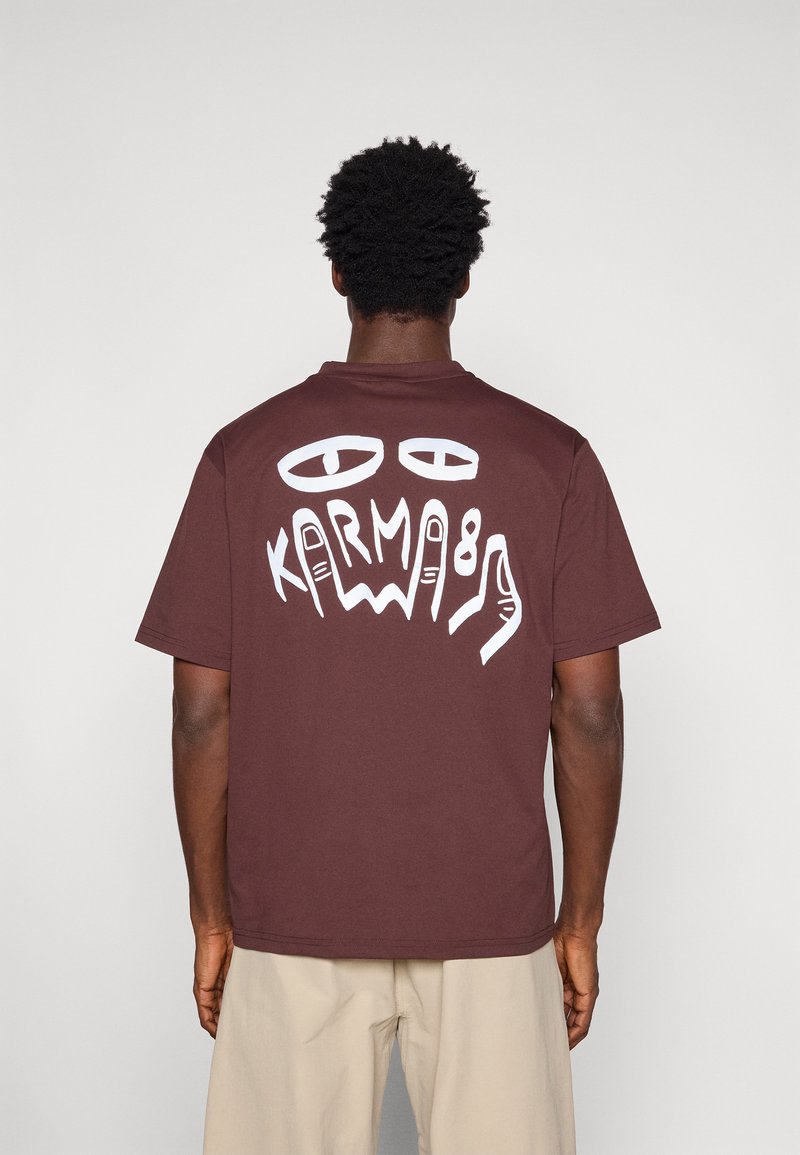 Uomo che indossa una maglietta marrone con occhi astratti bianchi e la grafica "KARMA 89" sul retro, pantaloni beige, in piedi contro uno sfondo semplice.