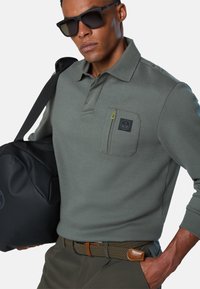 Polo verde a maniche lunghe con taschino e dettaglio della cerniera gialla. Indossato con una borsa nera, una cintura intrecciata e occhiali da sole.