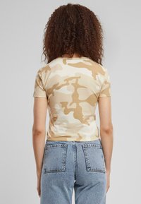 Lyse beige bomull t-skjorte med mørk beige kamuflasjemønster, kort erm og en tettsittende design. Kombinert med høyt liv lys blå jeans.