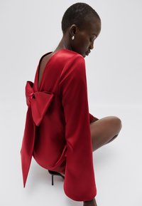 Red satin dress featuring a deep V-back, large bow detail at the back, and long, flowing sleeves. Model seated against a light background.