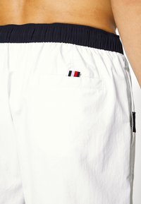 Pantaloncini da nuoto bianchi con una vita elastica blu navy, caratterizzati da una tasca posteriore e un piccolo logo a strisce nei colori rosso, bianco e blu.