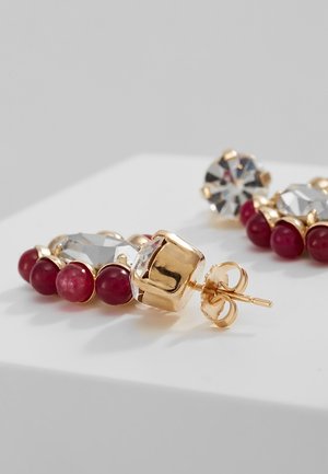 Gouden stekerearrings met heldere geslepen stenen en een halve cirkel van kleine ronde dieprode kralen op een wit oppervlak.