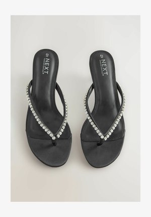 Fekete papucs sima textúrával, átlósan keresztbe futó pánttal, ami átlátszó gyöngyökkel van díszítve. Kényelmes viselet, egyszerű vonalvezetéssel tervezve.