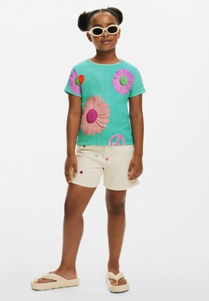 Türkisfarbene T-Shirt mit großen Blumenapplikationen, kombiniert mit beigen Shorts, die kleine bestickte Details aufweisen. Die Schuhe sind cremefarbene Sandalen.