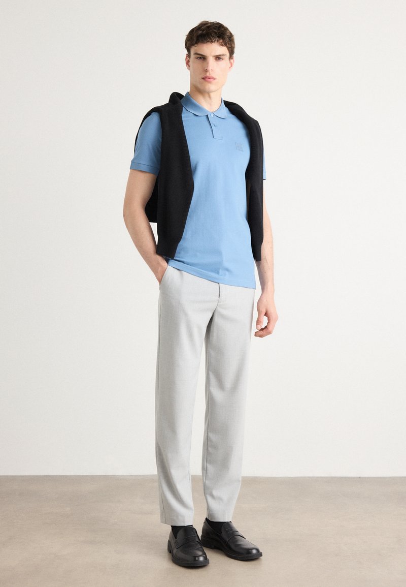 Polo de color azul claro, suéter negro colgado sobre los hombros, pantalones gris claro y zapatos negros. Accesorios mínimos, diseño limpio.