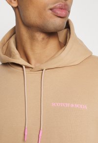 Felpa con cappuccio beige con coulisse, con logo rosa "SCOTCH & SODA" sul petto e una texture morbida.