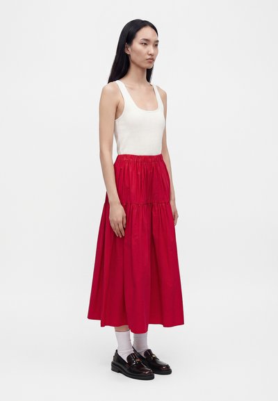 Femme aux longs cheveux noirs portant un haut blanc sans manches, une jupe midi rouge, des chaussettes blanches et des mocassins noirs, debout devant un fond uni.