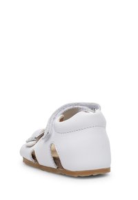 Zapato de cuero blanco con detalles recortados, punta redonda y correa ajustable. Suela plana con fondo marrón claro. Textura limpia y suave.