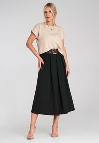 Beige Cropped-Top mit kurzen Ärmeln, kombiniert mit einem hochgeschnittenen, schwarzen Plisseerock, der mit einem breiten braunen Gürtel gesichert ist. Transparente Absätze runden das Outfit ab.