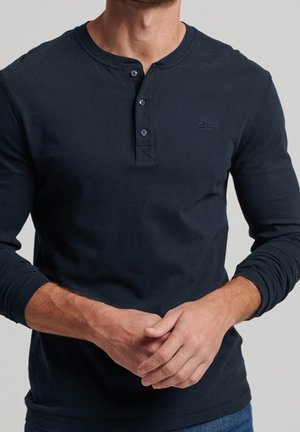 Mężczyzna ubrany w ciemnogranatową koszulę henley z długim rękawem i trzema guzikami, ręce złączone z przodu, widoczny od szyi do bioder.