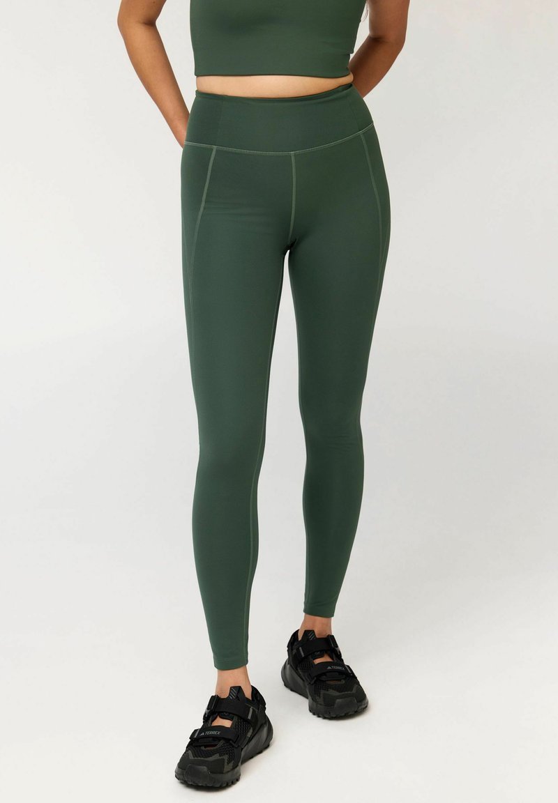 Vrouw die donkere groene leggings met hoge taille draagt, een bijpassende crop top en zwarte sportschoenen, staand met haar handen achter haar rug.