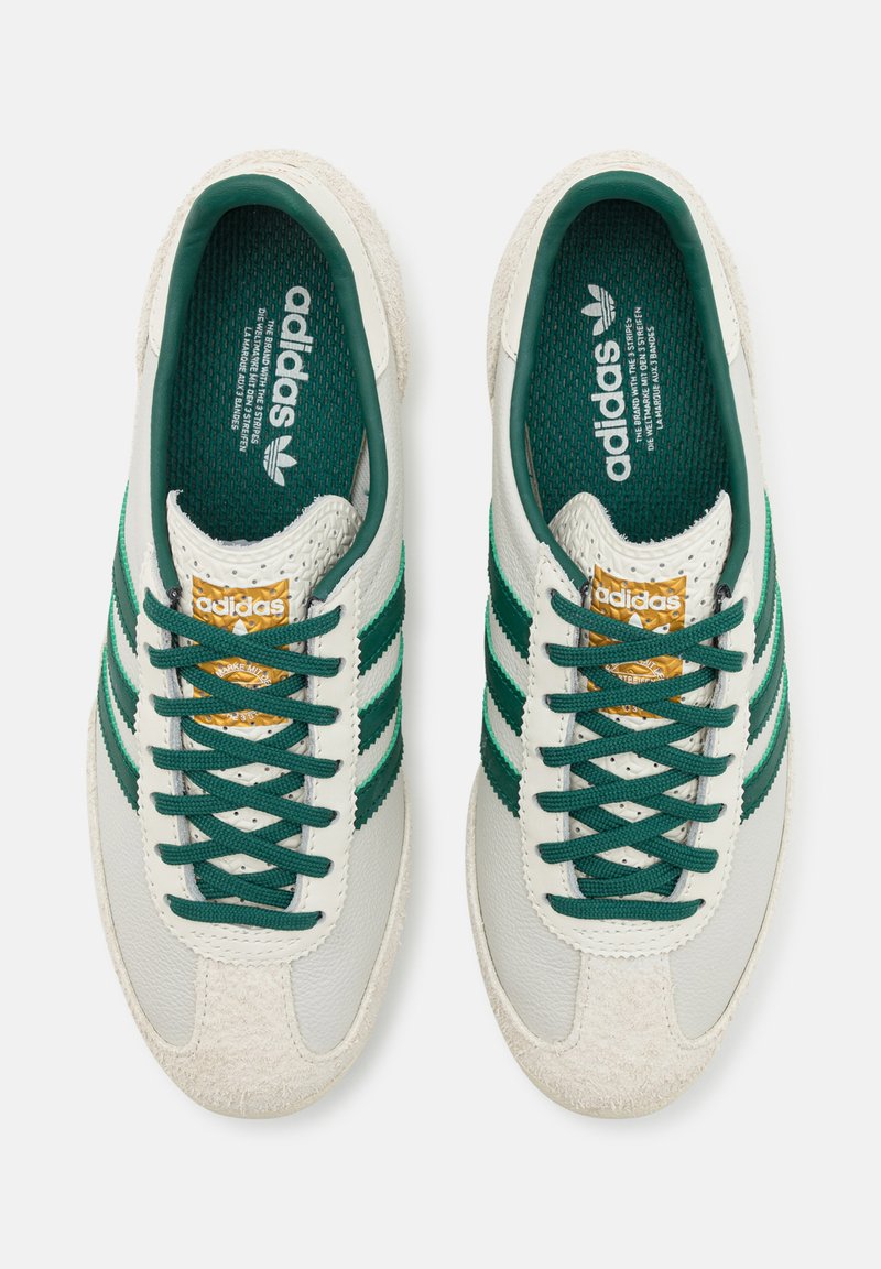 Baskets blanches en cuir avec des rayures et des lacets verts. Présentent un embout texturé et une étiquette dorée avec le logo de la marque.