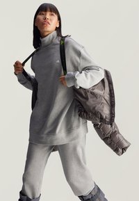 Szary sweatshirt z wysokim kołnierzem, pasujące szare spodnie dresowe oraz brązowa kurtka puchowa z siateczkowymi detalami. Torba z markowymi paskami.