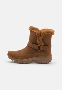 Bottine en suede marron avec bordure en fausse fourrure et une sangle étroite. Dotée d'une semelle en caoutchouc texturé et flexible et d'un design à bout arrondi.