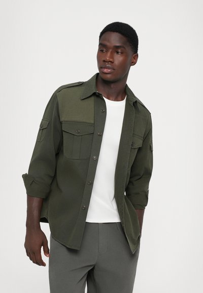 Olivgrüne Hemdjacke mit zwei vorderen Taschen, Knopfverschluss und hochgekrempelten Ärmeln, getragen über einem weißen T-Shirt und grauen Hosen.