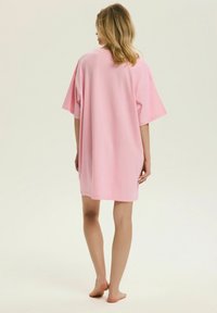 Kobieta stojąca boso, zwrócona tyłem, ubrana w luźną, różową sukienkę z krótkim rękawem sięgającą do kolan, na jednolitym tle.