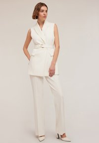 Chaleco blanco ajustado con cintura ceñida y bolsillos laterales, combinado con pantalones blancos rectos y tacones de punta. Acabado de tela suave.