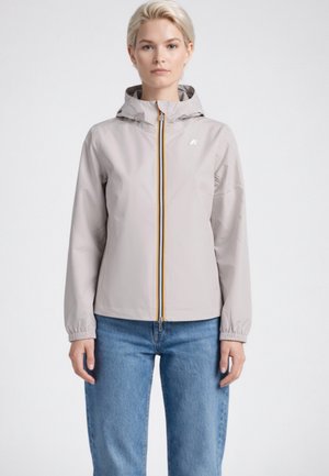 Donna che indossa una giacca con cappuccio grigio chiaro con cerniera e polsini elasticizzati, e jeans blu, in piedi davanti a uno sfondo bianco uniforme.