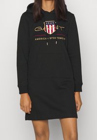 Robe sweat à capuche noire avec poche avant, featuring le logo "GANT" en design écusson doré, rouge et blanc, et accents de cordon de serrage.