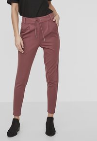 Pantalon pour femme bordeaux, ajusté, avec une taille à cordon, des plis à l'avant et des poches latérales, fabriqué à partir d'un tissu extensible. Bottines noires aux chevilles portées.